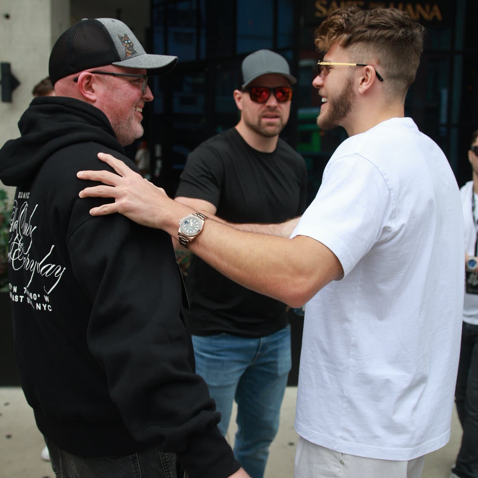 LambMetaX (@LambMetaX) in the center of a sidewalk conversation, grey cap and sunglasses, with two other people